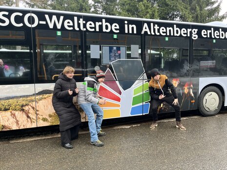 Drei Junge Personen posieren vor einem Bus mit dem Stollnmundlochlogo des Welterbevereins Montanregion Erzgebirge e. V.